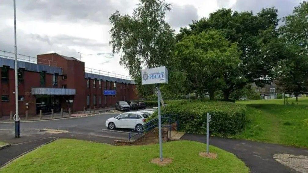 The front of a police station. In the foreground are grass verges, a sign saying West Yorkshire Police and parked cars. The station itself is a red-brick building set back from the road.