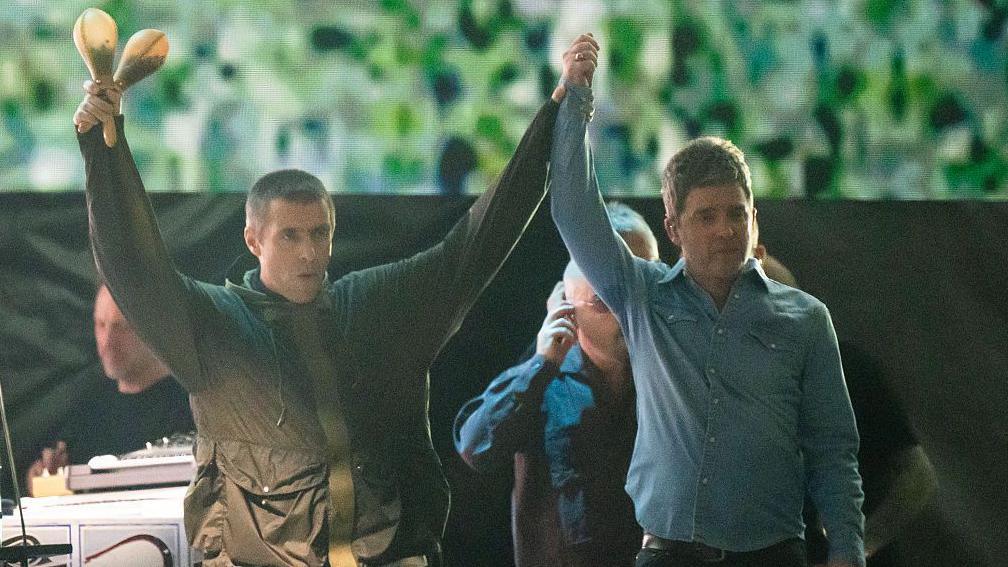 Gallagher brothers Liam and Noel arrive hand in hand on Principality Stadium. Noel wears a denim shirt while Liam wears a green parka. 