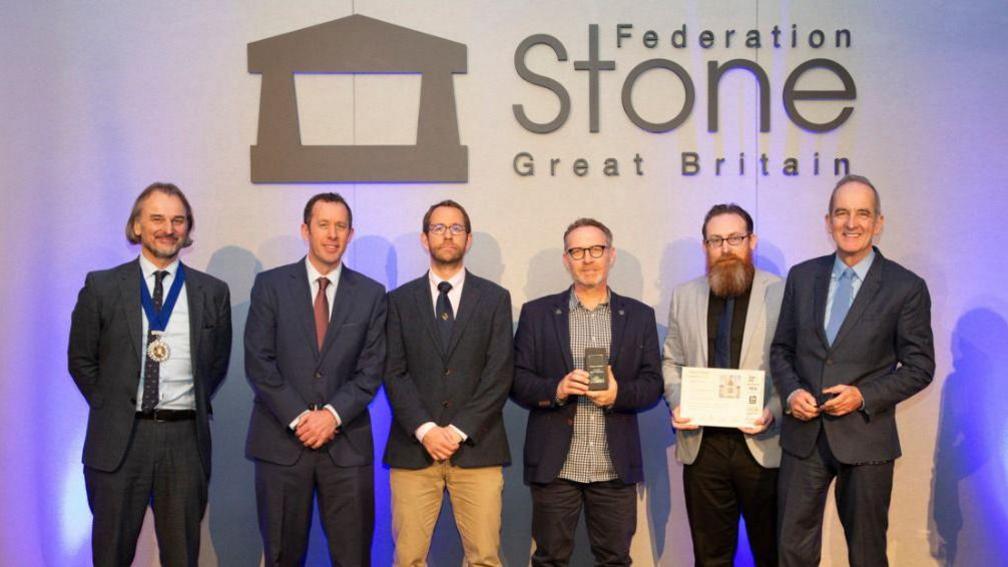 Six middle-aged men dressed smartly in suits, one holding an engraved award and another, a certificate, in front of a wall with 'Federation Stone, Great Britain' written on it. 