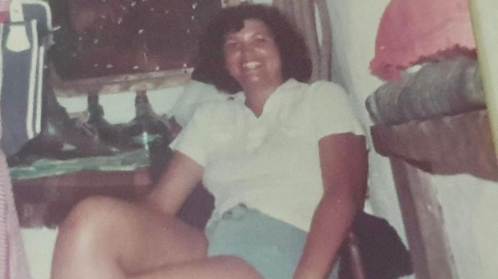 Lady with a brown short perm, wearing a white shirt and blue shorts