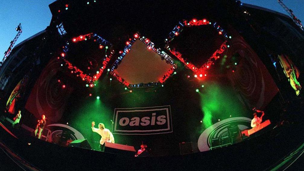 Oasis on stage. Green stage lights illuminate them both, with a black backdrop with the Oasis logo on it.