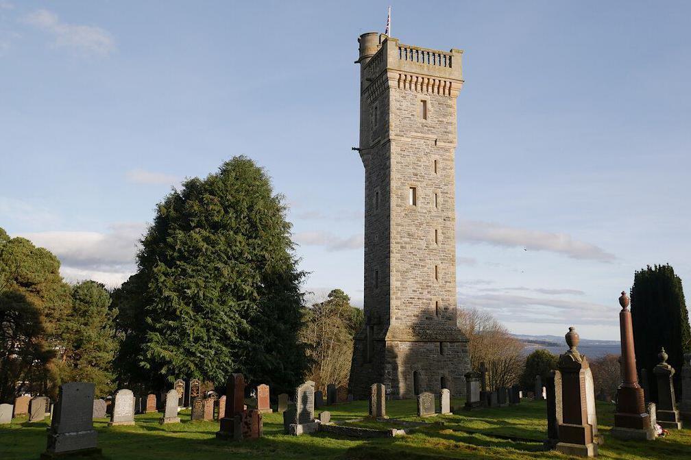 The stone memorial towers above a cemetery and trees. Sunlight strikes one side of the tower. The sky is blue with a few clouds.