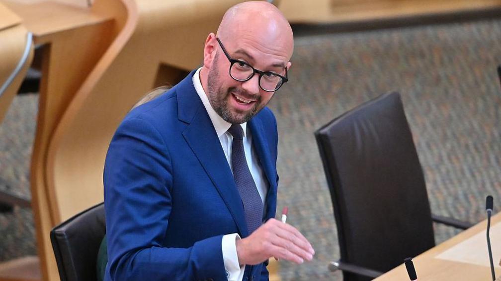 Ben Macpherson speaking in the Scottish Parliament, while gesturing with his right hand. He is a bald man with glasses and a beard and is wearing a blue suit, white shirt and a tie.