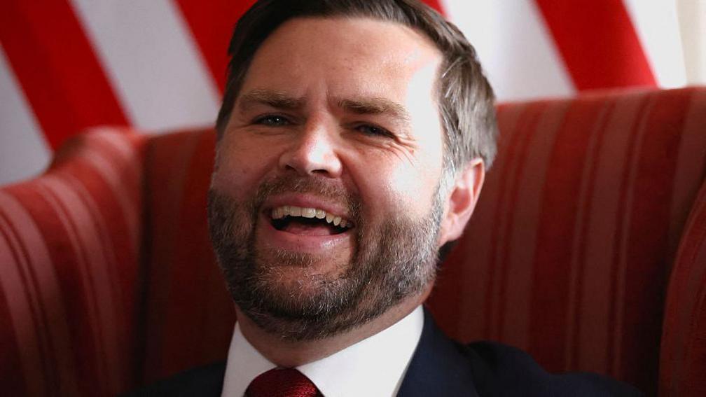 Close up image of JD Vance, laughing, with a US flag behind him