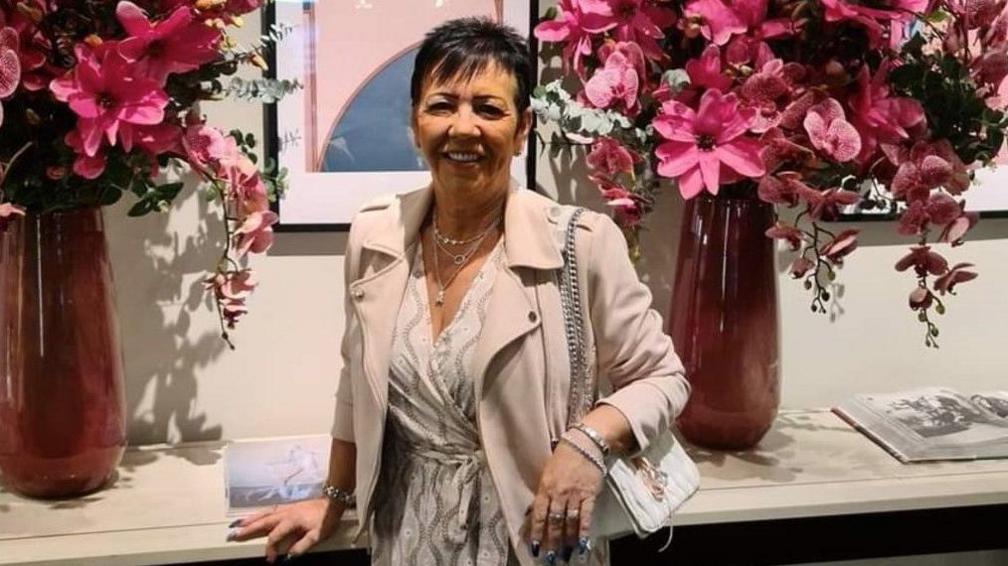 A middle aged woman is facing the camera smiling.  She is wearing a cream coloured jacket with a cream top underneath.  She is standing in front of a shelf with vases of bouquets of pink and white flowers On either side of her.