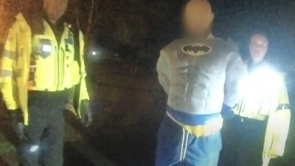 A man wearing a Batman costume with his hands behind his back stands next to two police officers