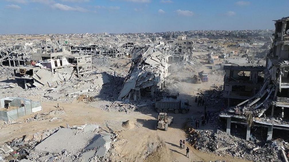 A picture from 28 October, 2025 shows a truck and four other vehicles surrounded by rubble in Khan Yunis. All the buildings in the foreground and middle-distance have been totally collapsed into dust and debris.