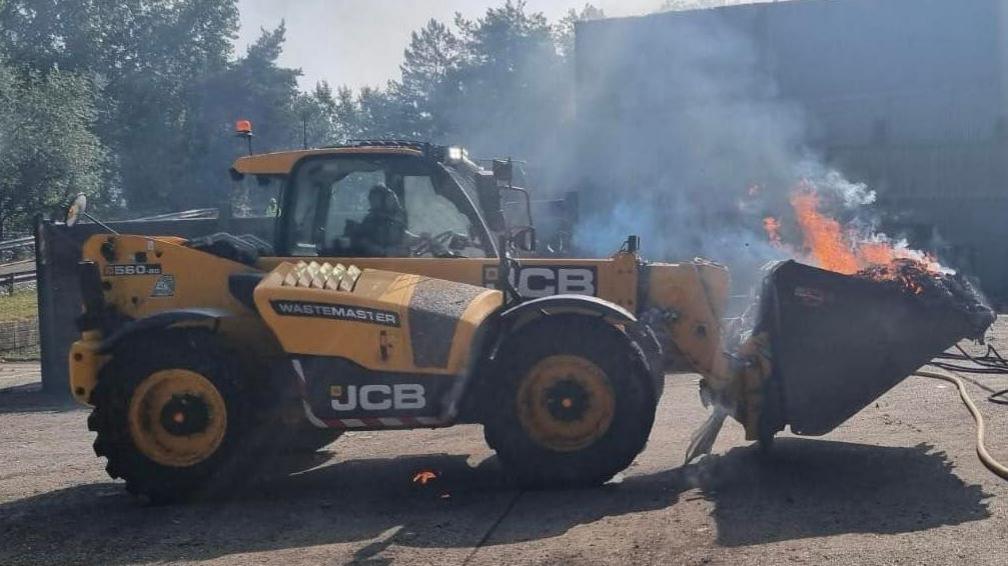 A yellow and black JCB telehandler with burning waste in its bucket