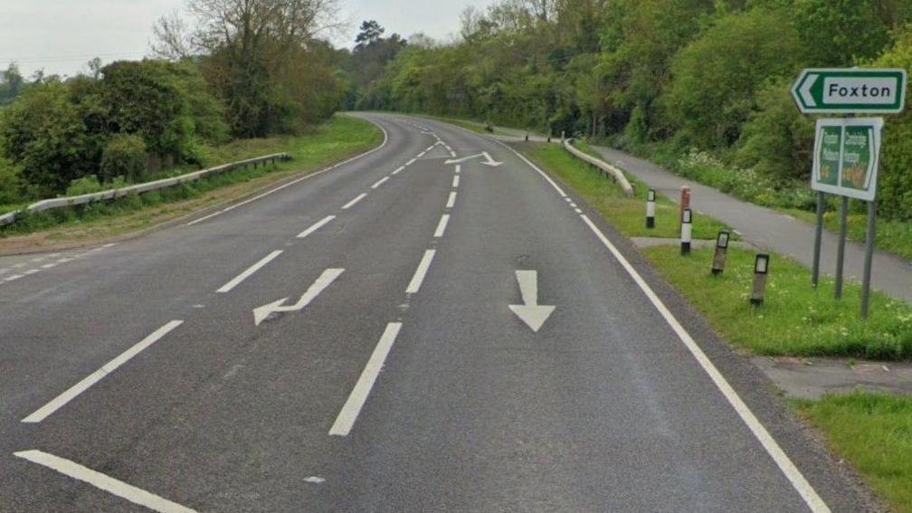 The A10, a dual-carriage way, with a road sign to the left, with arrows on the road, and a chevron in the middle. There is grass on either side of the road, and trees. 