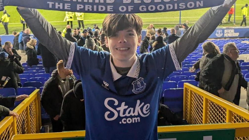 Mackenzie grins as he holds a blue Everton scarf aloft saying "The Pride of Goodison".