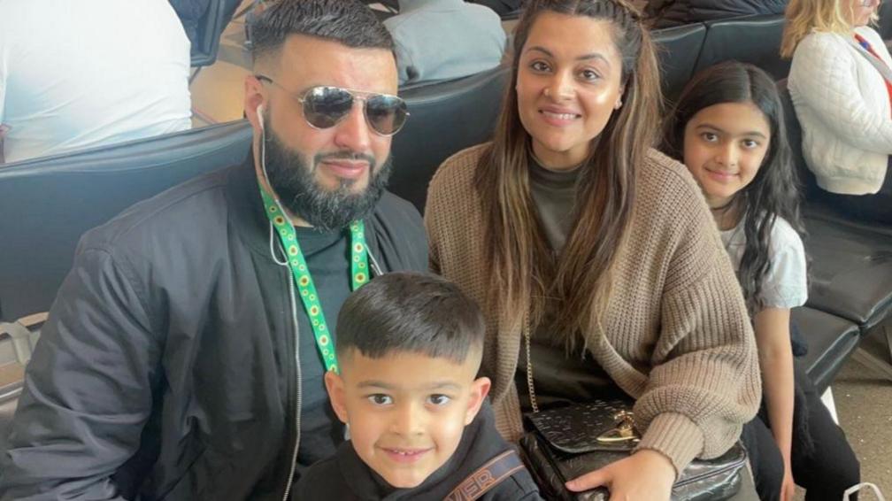 A young family cuddling up together for a photo. There is a man and woman and two children.