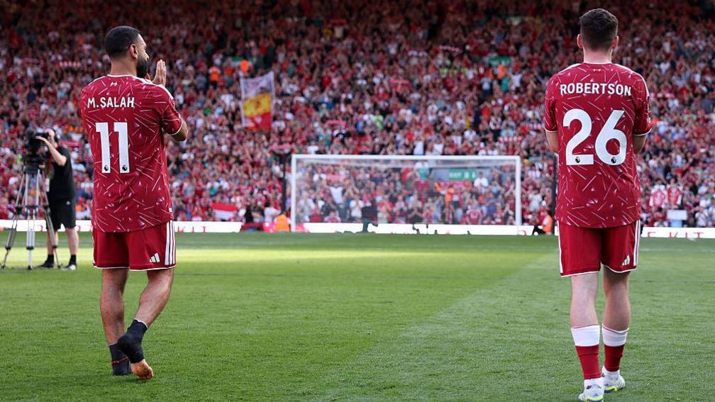 Mohamed Salah and Andy Robertson, in red Liverpool kit with their names on the back, have their backs to the camera as they look towards supporters on the club's Kop stand