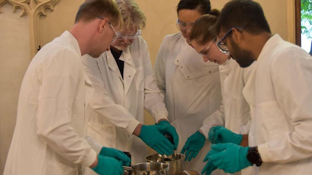 Prof John Carr (second left) with Dr Betty Chung (back left) and Robbie Waddell (front right), Jennifer Palmer (second right) and Satish Viswanathan (far right) all in white lab coats and clear safety goggles in Corpus Christi College chapel