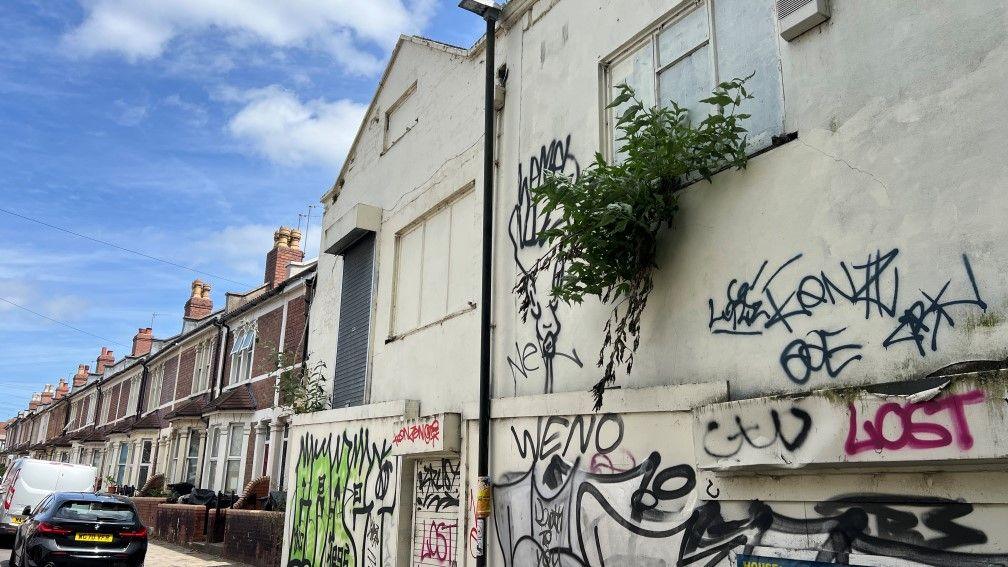 An empty property in St Werburgh's, Bristol. The building is covered in graffiti and there is a row of terraced houses beside it.