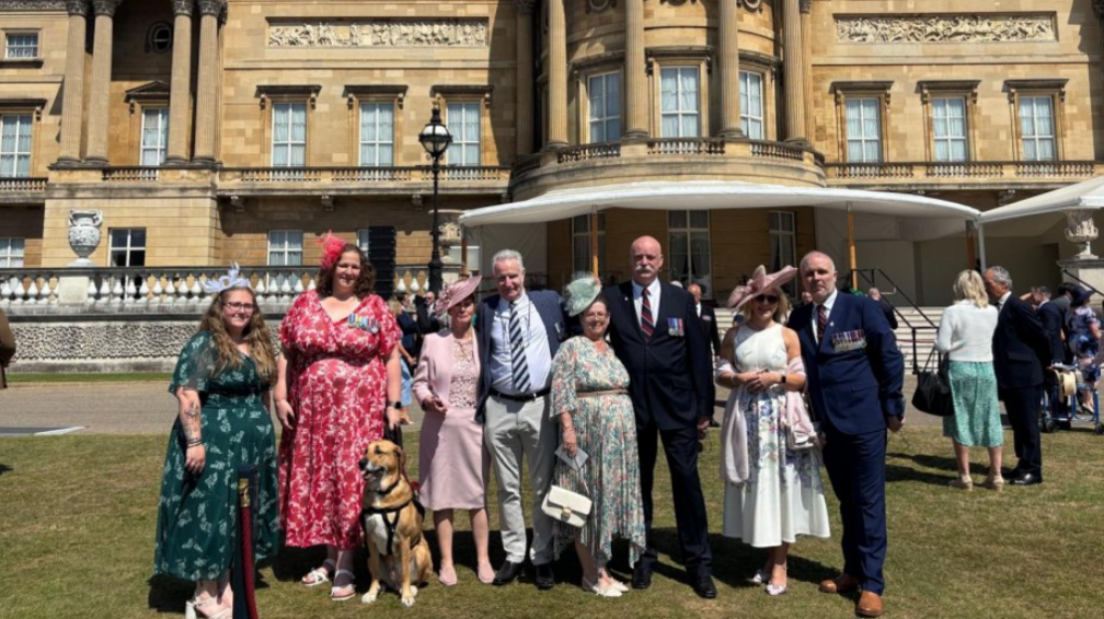 A group of men and women in smart clothing standing on a lawn in front of Buckingham Palace which has columns and ornate decorations with large sash windows. The women are wearing dresses with hats and the men are wearing suits. There is also a tan coloured dog with the group of people who are all smiling.