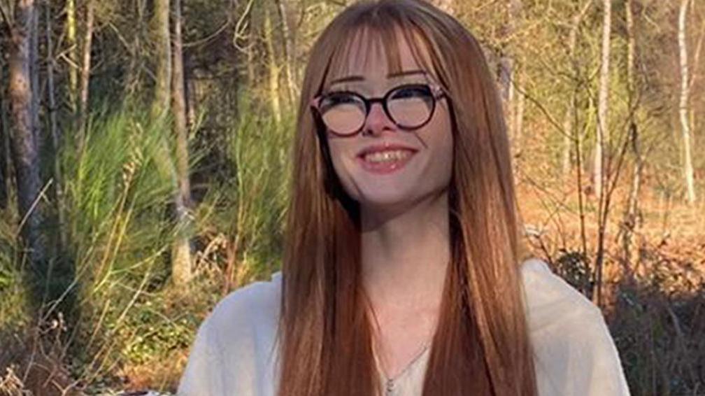 Brianna Ghey, with long auburn hair and round glasses, smiles and wears a white zip-up top in front of autumnal woodland and snowy ground