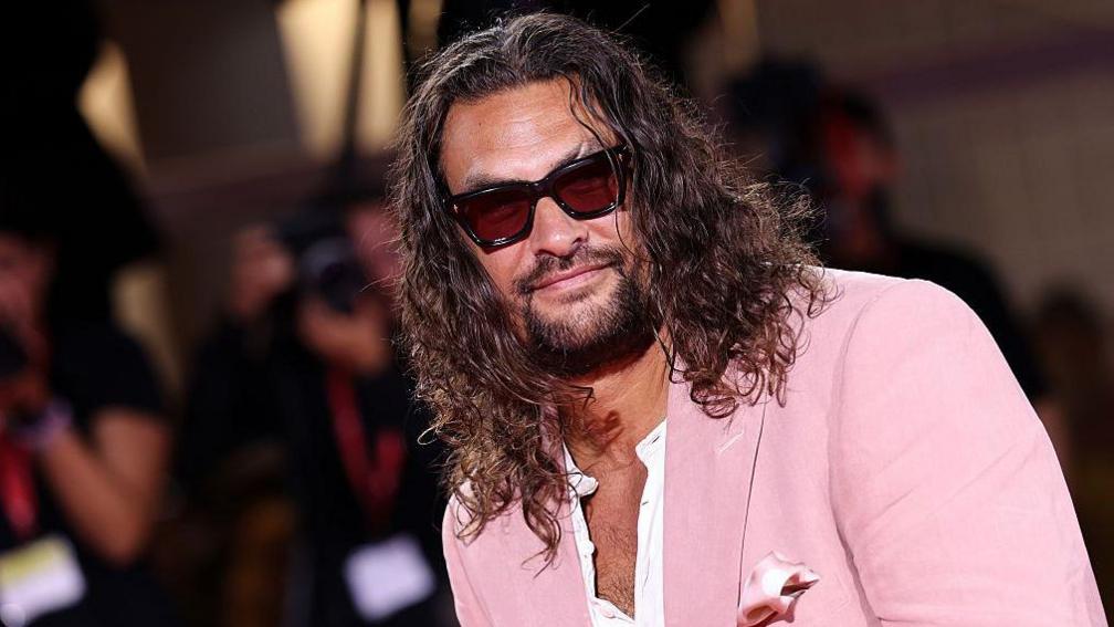 Jason Momoa attending the Cartier Glory To The Filmmaker Award 2025 and the "In the Hand of Dante" red carpet during the 82nd Venice International Film Festival. He has long, wavy dark brown hair and a dark beard. He is wearing a pink suit jacket over a white shirt which is partially unbuttoned and black rimmed sunglasses. The blurred image of photographers can be seen behind him.