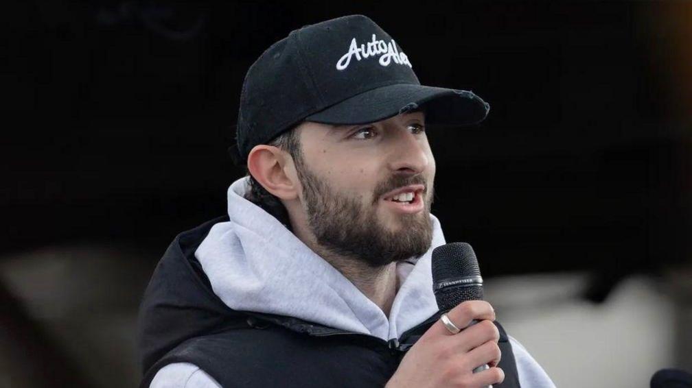 A man wearing a hoodie and a black puffer jacket. he is also wearing a cap that reads AutoAlex and is talking into a microphone.
