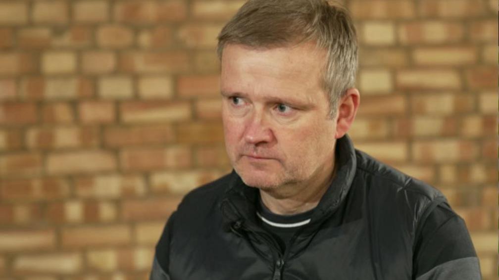 Mr Turner wears a black jacket and sits in front of a brick wall. He has short hair and no facial hair.