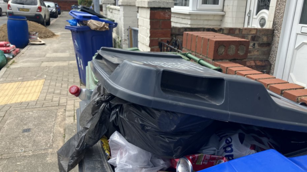 An overflowing blue wheelie bin and an overflowing black wheelie bin on a street with terraced houses and cars parked on the road. 