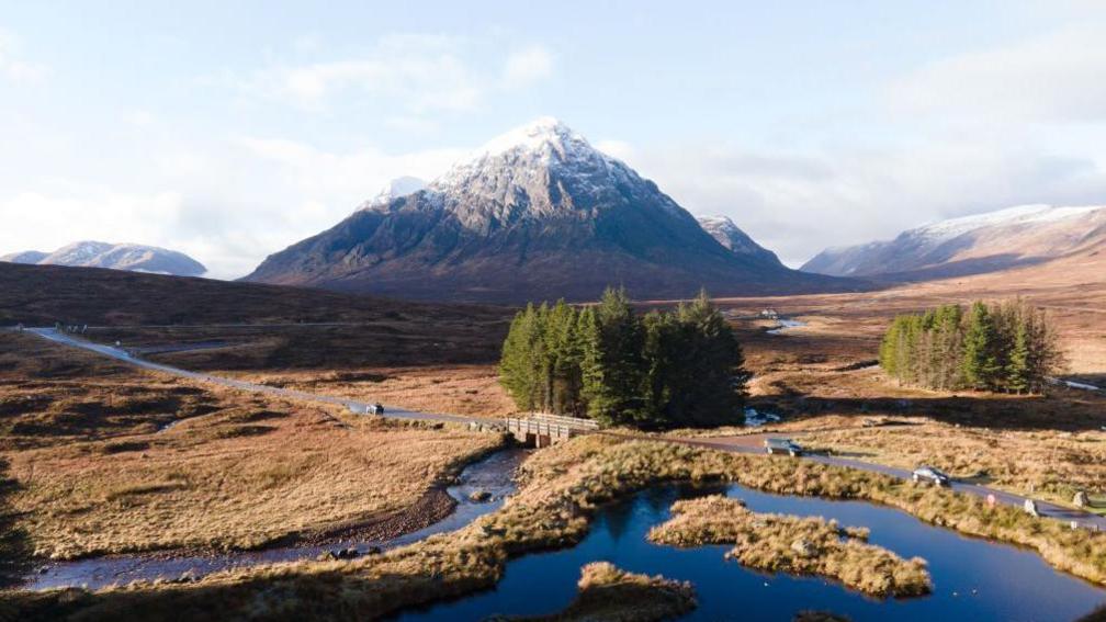 A rural road runs across the Highlands with a snow-capped mountain in the distance