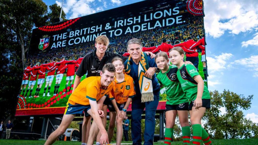 Tim Horan and some children stand in front of advertising hoarding with the Lions