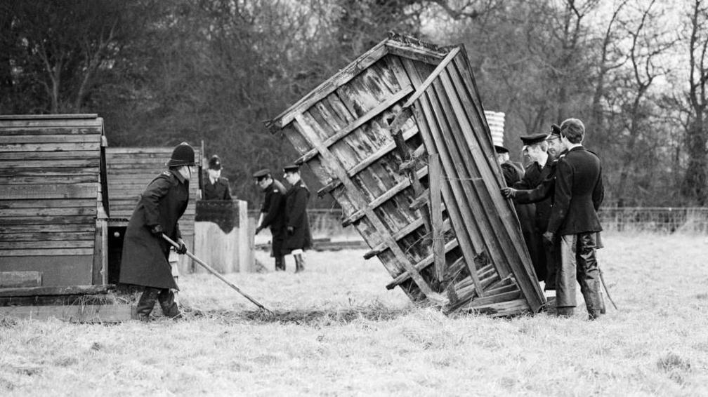 Black and white image of police searching Rooks Farm, and holding up a pig hut