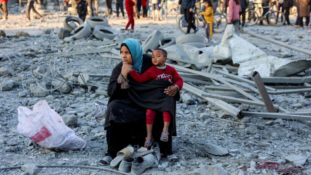 A woman, sitting in rubble in Gaza, with a boy on her lap. There are also people in the background. 