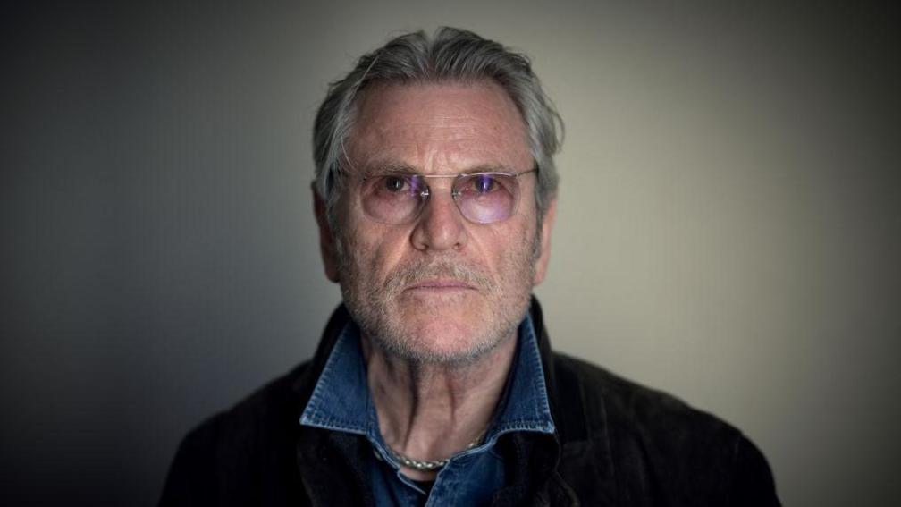 A photo of Tchéky Karyo, taken at La Baule film festival in July 2022 - he has grey hair, a beard and wears glasses and a denim shirt.