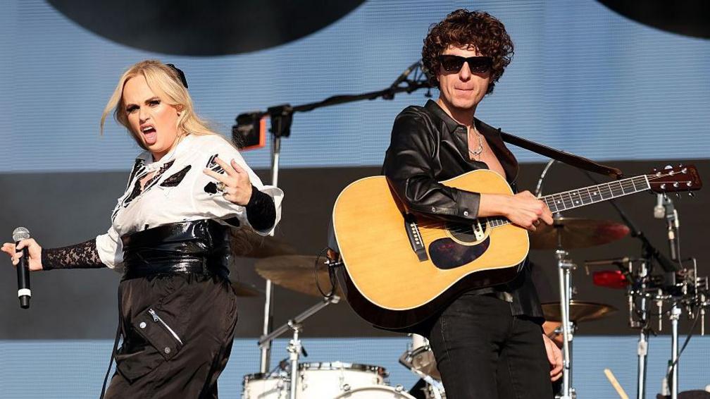 Actress Rebel Wilson, wearing cargo pants and a white shite, makes a surprise appearance on stage with The Kooks' lead singer Luke Pritchard, who is wearing a leather jacket and sunglasses. He is holding an acoustic guitar across his body. 