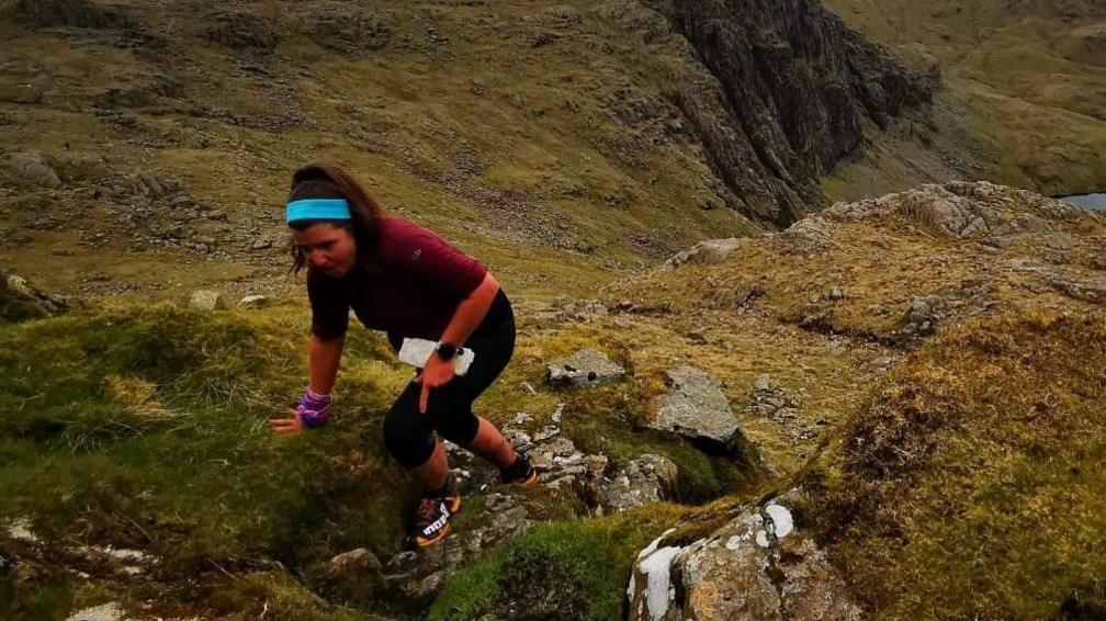 Fran Blackett is climbing up a hill clutching a map, with her hand on the grassy moorland. 