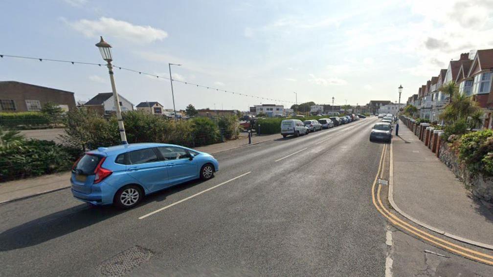 A Google Maps image of a junction. There is one blue car driving on the road, and others parked up on either side. 