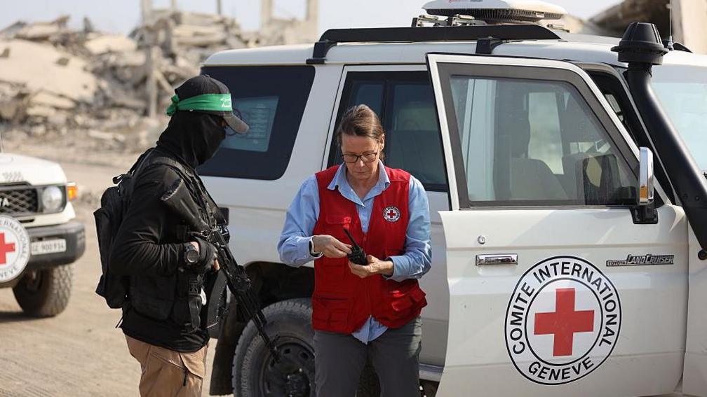 A Red Cross worker stands outside the open right-hand side door of one of the white Red Cross cars, wearing a blue, shirt and a red vest with the Red Cross logo on the left-hand side, turns a knob on a radio communication device, watched by an individual carrying a rifle across his chest, with beige trousers and black jacket, as well as a black balaclava and hat, which has the green Hamas bandana around
