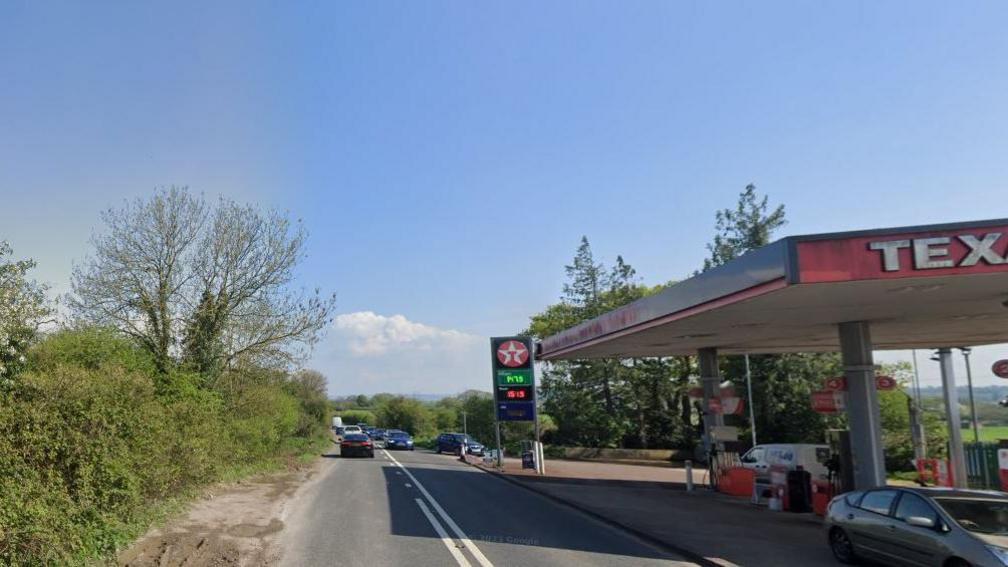 A rural two-lane road with hedgerows on each side, a Texaco garage sits on the right hand side of the image. 