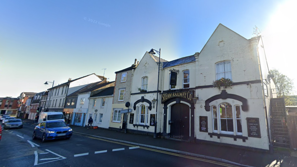 A Google Maps streetview picture of the Steam Railway pub. The building is double fronted with two pointed roofs, rendered in cream plaster with a black sign over the front door which reads 'Steam Railways Co' in gold lettering.  