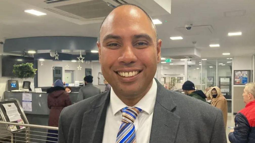 Dr Neil Modha in a GP surgery smiling at the camera. He has short hair and is wearing a suit and tie. Behind him are patients queuing for the reception.