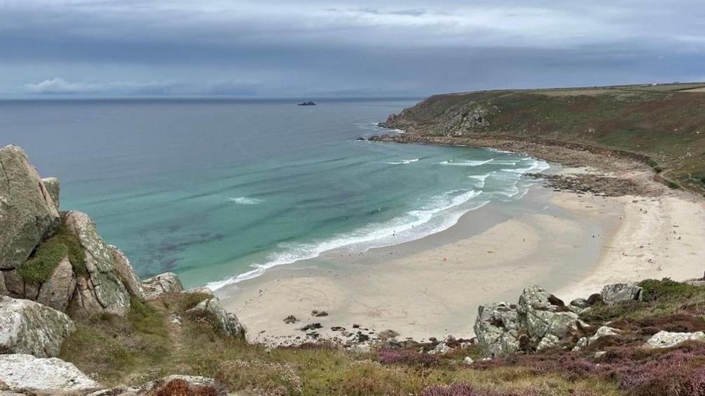 Gwynver Beach at Sennen, a large sandy beach with headland on either side. The image is taken from the top of a cliff looking down over the beach. The water is blue.