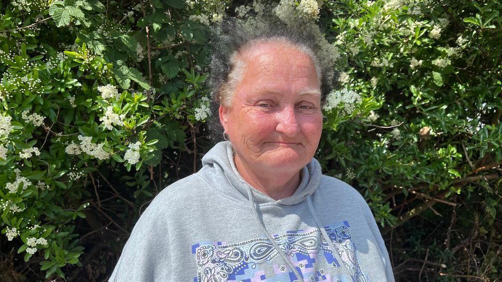 Woman in grey fleece with pattern on stands smiling with green hedge behind