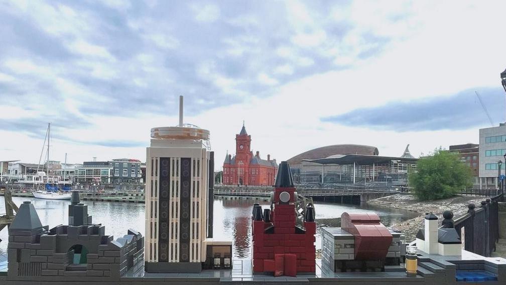 A Lego city-scape, with buildings including Cardiff castle and the Pierhead building lined up in a row, with the real Cardiff Bay in the background