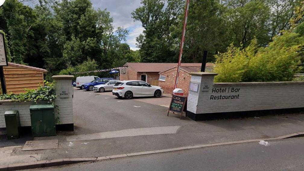 A sign including the words Cask and Tandoor is placed next to a white wall by the pavement by a road. Cars are parked in the background of the photo.