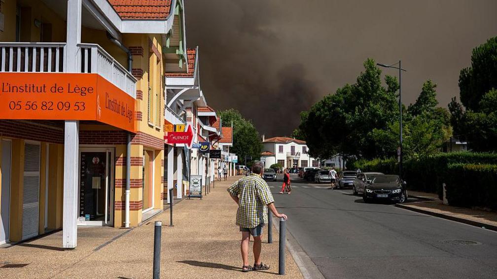 Pedestrians look at a cloud of black smoke covering the sky nearby of an area evacuated due to a wildfire in Lege Cap-Ferret.