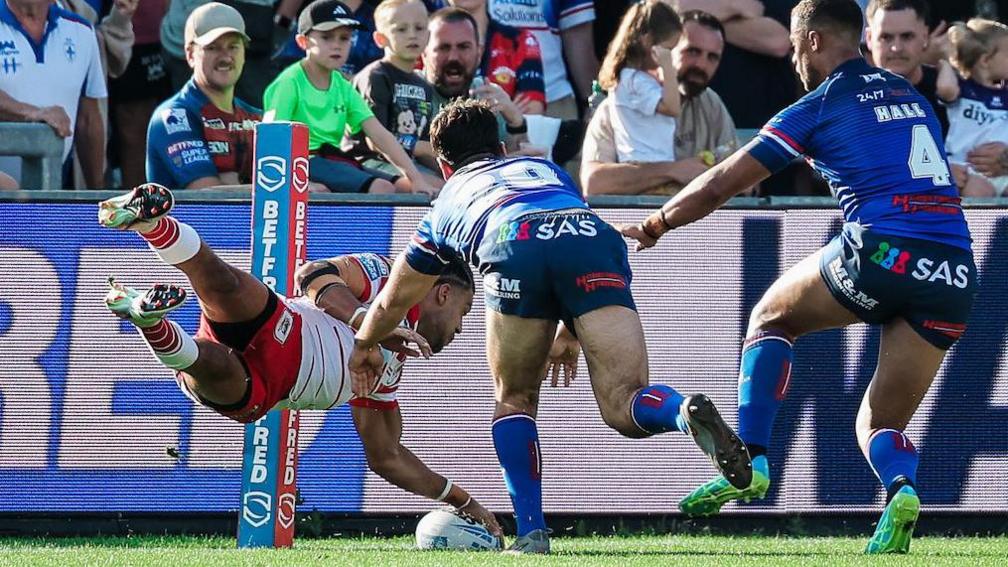 St Helens wing Lewis Murphy scoring a try for his side by diving for the line with his left hand on top of the ball which is grounded on the line while two Wakefield defenders try to stop him