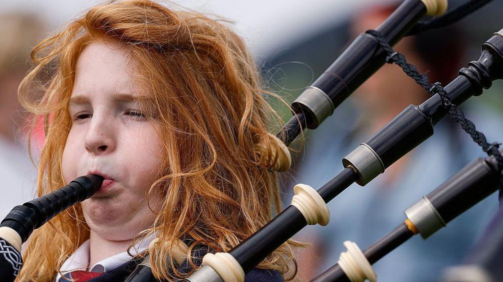 Close-up photograph of a teenager playing bagpipes. The piper has long, wavy, red hair and their cheeks are puffed out as they blow into the mouthpiece