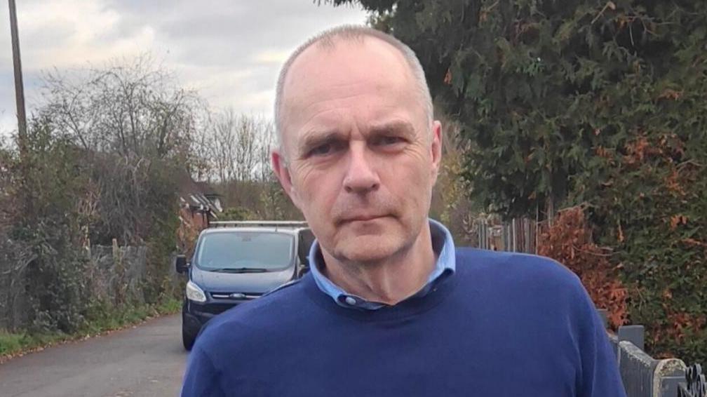 A man with short grey hair stood looking into the camera while beside a road, on his left. He is wearing a light blue shirt and dark blue jumper. He has a blank expression on his face/