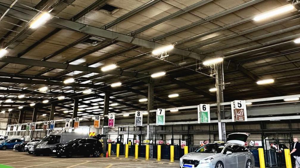 Inside the recycling centre under a metal roof, with cars parked in bays. There are number cards for bays placed above the vehicles.