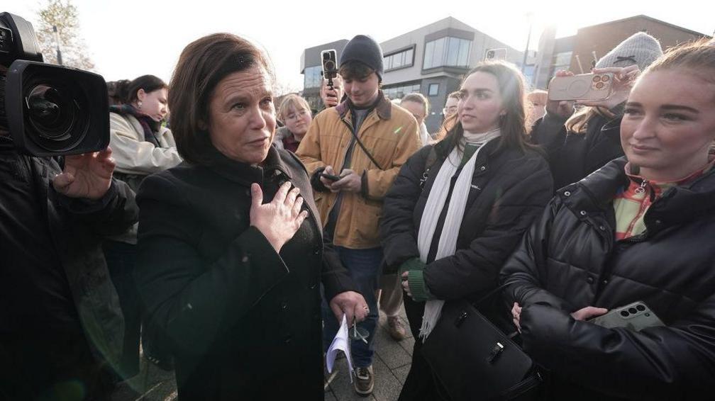 Mary Lou McDonald with shoulder length dark hair holds her hand to her chest as she talks to two female students both wearing dark coats at an outdoor event in Dublin