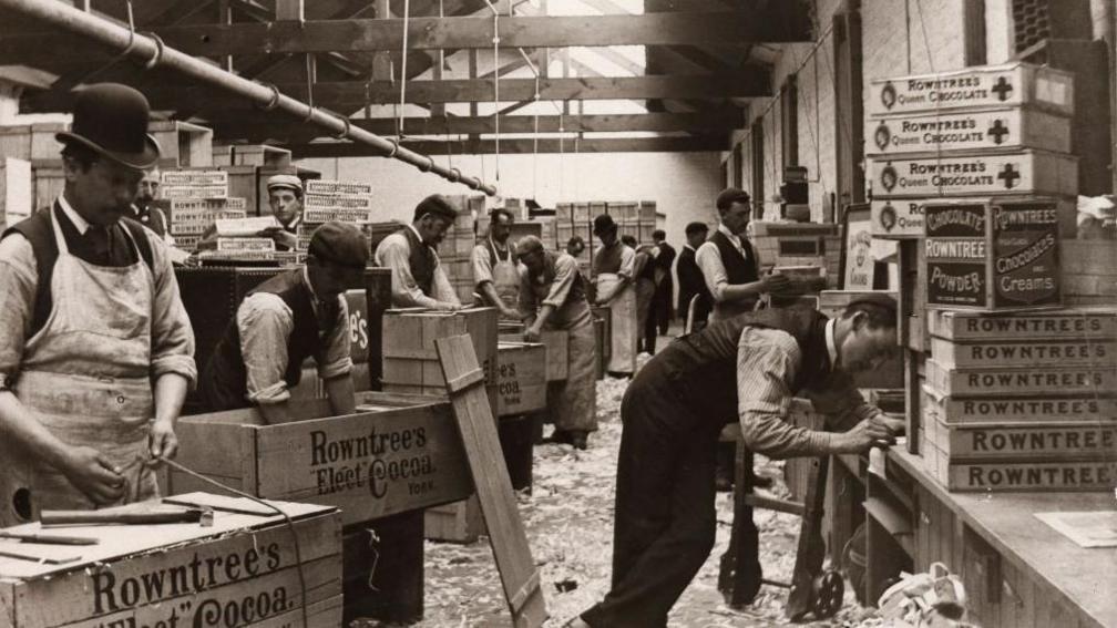 A black and white photograph showing a warehouse with men wearing white aprons and working over wooden boxes. The boxes say Rowntree's Elect Cocoa on them. Several of the men are wearing black bowler hats while they work and their shirt sleeves are rolled up. On a desk along the wall, boxes are piled up. Some say Rowntree's Queen Chocolates on them, other piles say Rowntree's on the boxes or Rowntree's Chocolate Powder.