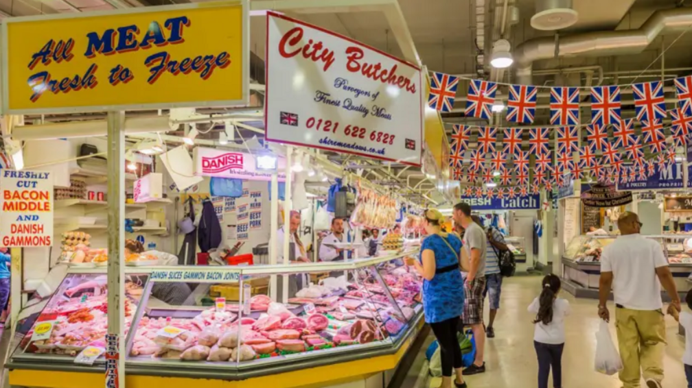 People look at a colourful butcher's market stall. A sign above the stall says all meat fresh to freeze and another says "city butchers".