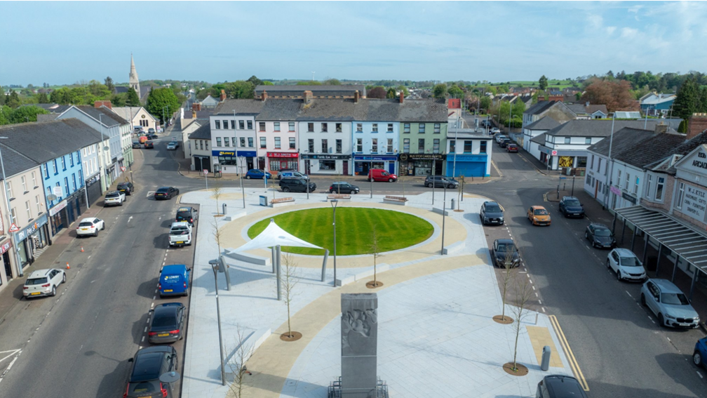 Scene from the centre of Castlederg, with grassy circle in the centre. There is a stone sculpture at the front. The centrepiece is surrounded by cars and houses.