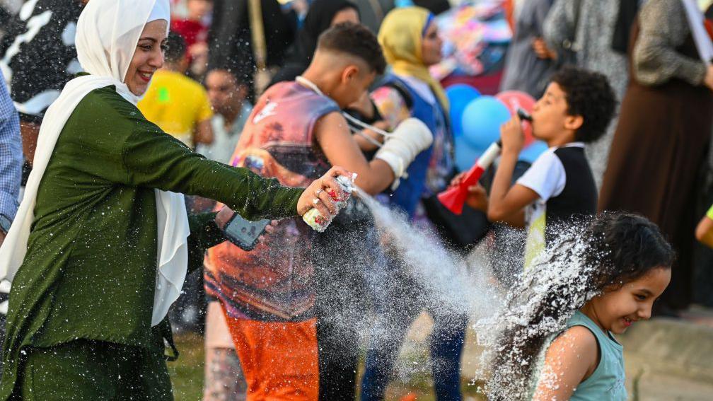 A woman playfully sprays a girl with foam as they celebrate Eid